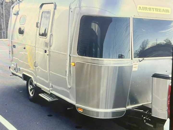 Shiny Airstream rv rental travel trailer parked on asphalt. Its polished silver exterior reflects trees and sky, offering a modern look.. Airstream Caravel 2020