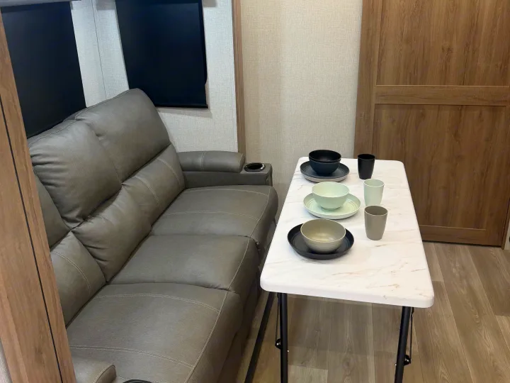 An inviting RV rental space featuring a comfortable gray sofa, a dining table with modern dishware, and light wood accents.. Grand Design Imagine XLS 2024