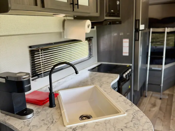 Inside a modern RV rental: sleek kitchen with sink, Keurig, gray cabinets & marble-look counters. Bunk beds in the background.. Dutchmen Aspen Trail LE 26BH 2022