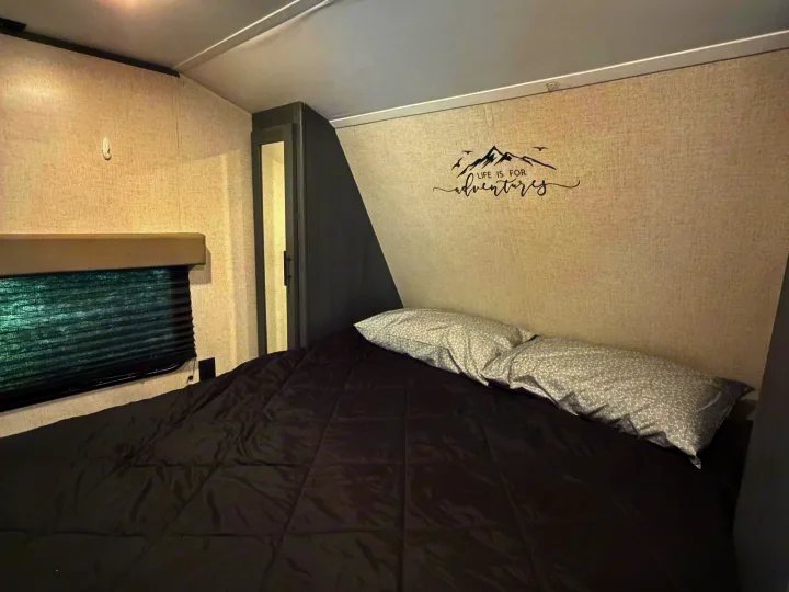 Inside an RV rental, this cozy bedroom features a black comforter, patterned pillows, and a "LIFE IS FOR adventures" wall decal.. Dutchmen Aspen Trail LE 26BH 2022
