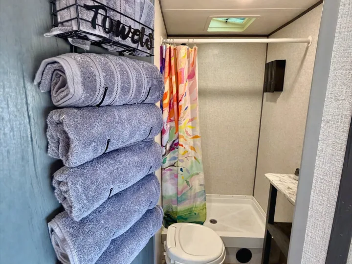 This RV rental bathroom shows a wall-mounted towel rack, colorful shower curtain, and toilet, offering practical use in a compact space.. Dutchmen Aspen Trail LE 26BH 2022
