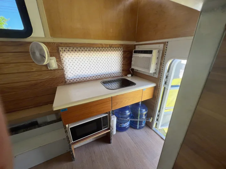 Compact RV rental interior: kitchenette with sink, microwave, AC, fan, and water bottles. Wood-paneled walls.. Ford Ford F150 2019