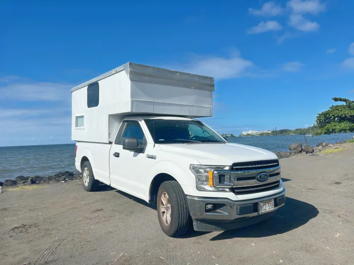 White Ford F-150 pickup with a custom RV rental camper unit, parked by a rocky ocean shore under blue skies.. Ford Ford F150 2019
