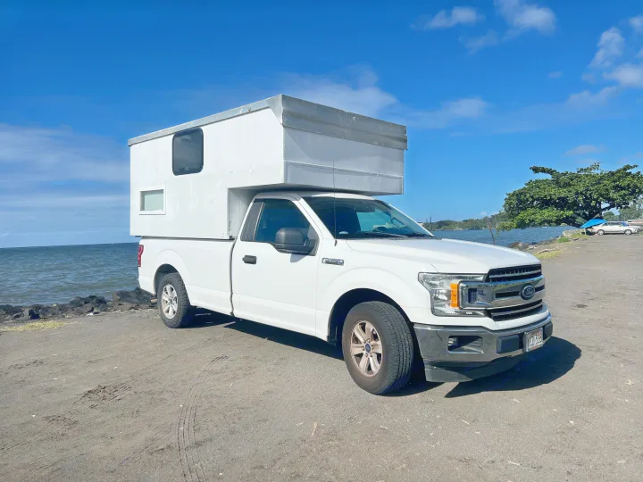 White Ford F-150 pickup with a truck bed camper, perfect for an RV rental, parked by the ocean on a bright day.. Ford Ford F150 2019