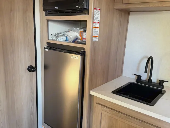 RV rental kitchen area features a microwave, mini-fridge, and sink on a white countertop, alongside a wooden door.. Keystone RV Coleman 2025