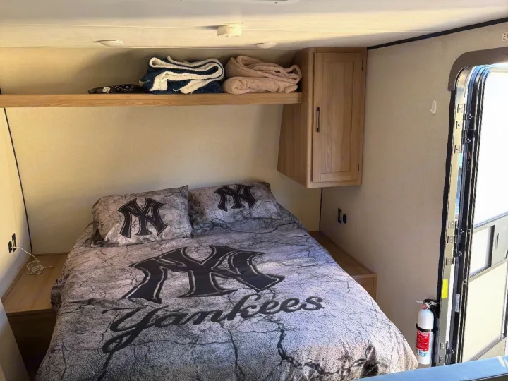 Cozy RV rental sleeping area with New York Yankees bedding, storage shelf, and cabinet. Fire extinguisher by the door.. Keystone RV Coleman 2025