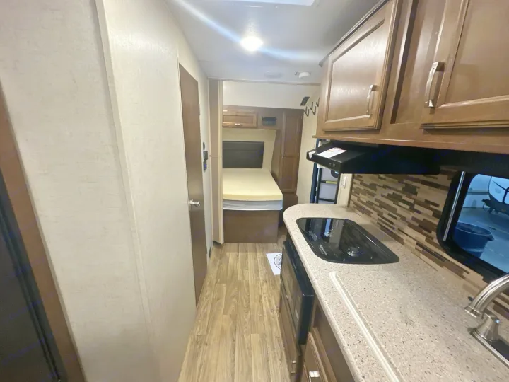 This RV rental interior features a compact kitchen with a stovetop and sink on the right, and a bedroom visible in the background.. Venture RV Sonic 170 VBH 2017
