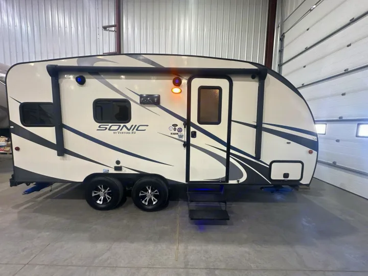 Exterior of a light-colored Sonic travel trailer, a potential rv rental, with a deployed awning and blue-lit entry steps.. Venture RV Sonic 170 VBH 2017