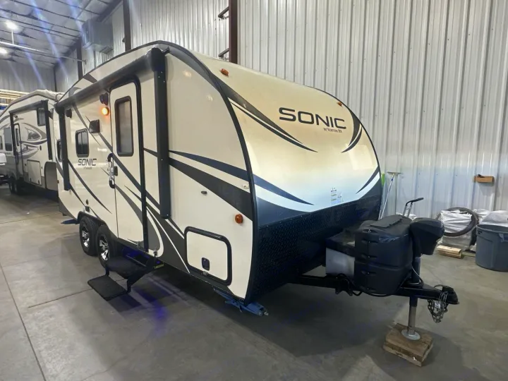 Cream and black Sonic by Venture RV travel trailer, perfect for an rv rental, parked indoors. Steps are extended.. Venture RV Sonic 170 VBH 2017