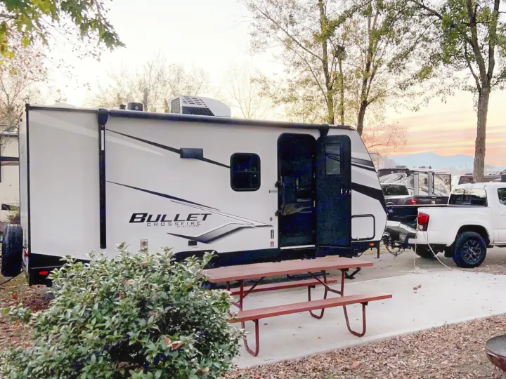 RV rental: A white travel trailer and pickup truck parked at a scenic campsite with a picnic table and fire pit. Sunset hues light the sky.. Keystone RV Bullet Crossfire 2024