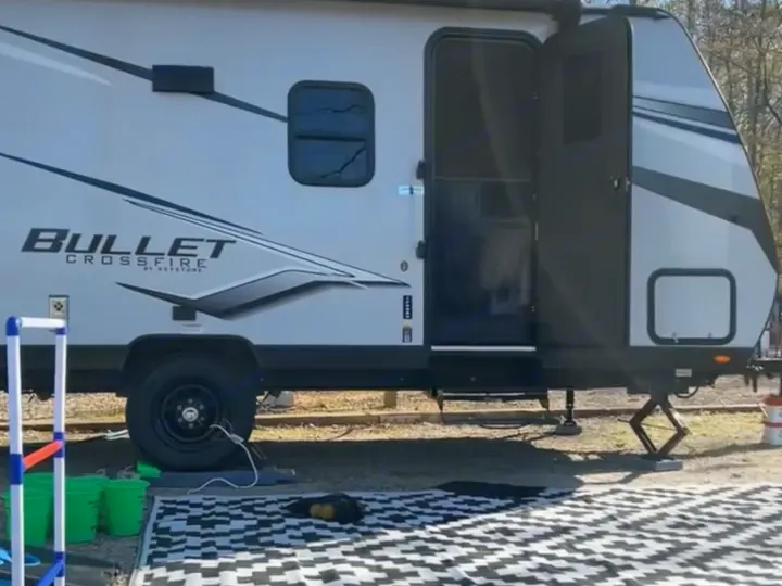 rv rental at a sunny campsite. A white travel trailer is parked with an outdoor rug and ladder toss game. Trees background.. Keystone RV Bullet Crossfire 2024