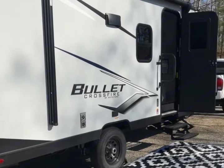 Enjoying an RV rental setup! A white Bullet Crossfire travel trailer with an open door, awning, and outdoor mat on a sunny campsite.. Keystone RV Bullet Crossfire 2024