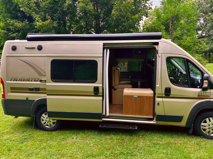 A modern Travato Promaster camper van, an ideal RV rental, parked on grass with its side open, showing interior seating and table.. Winnebago Travato 2014