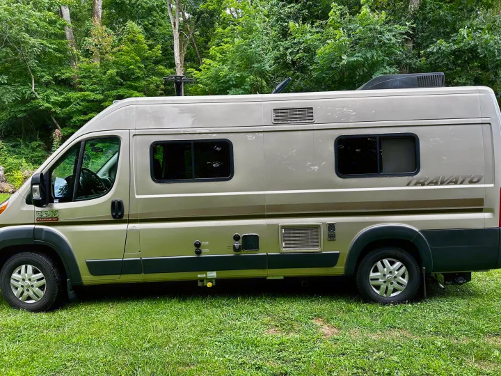 RV rental: Tan Ram ProMaster Travato camper van parked on green grass in a wooded area, showing its side profile.. Winnebago Travato 2014