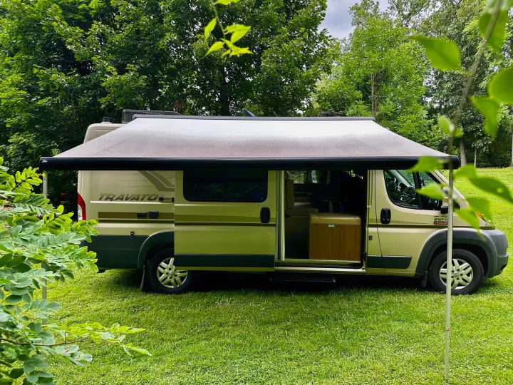 Gold RV rental camper van parked in a grassy, wooded campground. Its awning is extended, and the side door is open, inviting outdoor adventure.. Winnebago Travato 2014