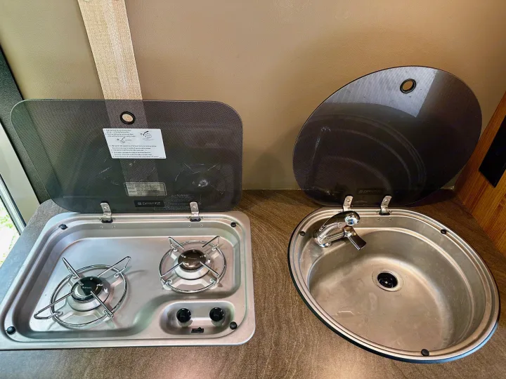 RV rental kitchen featuring a compact Dometic two-burner stove and sink with hinged glass covers open.. Winnebago Travato 2014