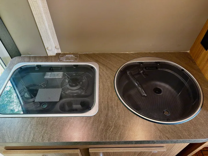 Compact RV rental kitchen featuring a Dometic stovetop and sink, both with closed lids, set into a patterned countertop.. Winnebago Travato 2014
