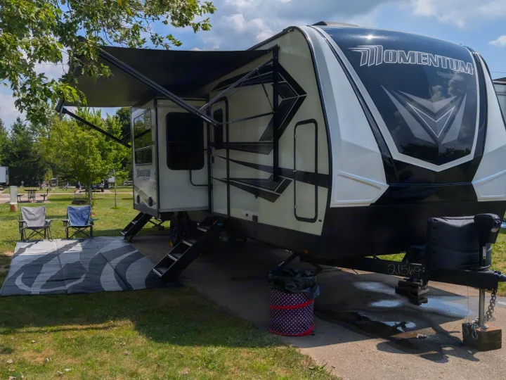 Spacious RV rental set up at a grassy campsite with awning, outdoor rug, and chairs. Perfect for a relaxing getaway.. Grand Design Momentum Toy Hauler 2022