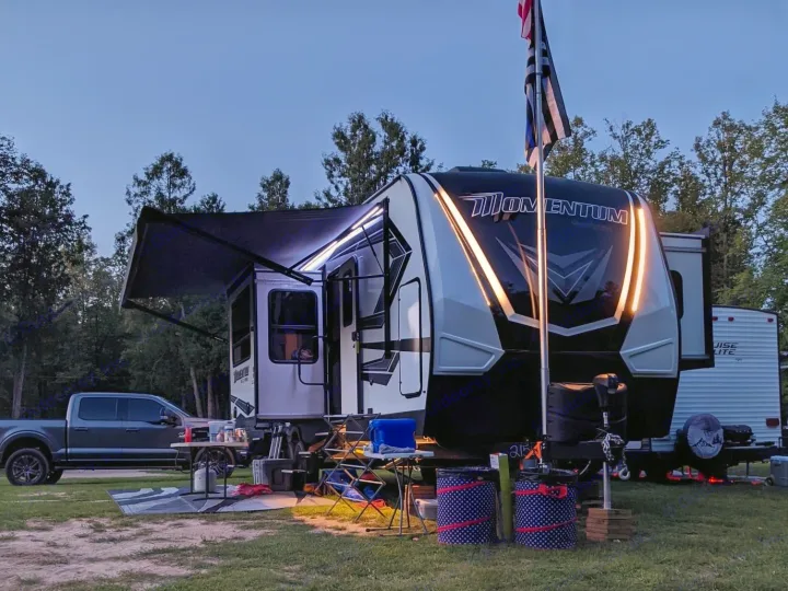 Evening RV rental setup: Large camper, truck, flags, and outdoor gear in a dark campground. Doll strollers visible.. Grand Design Momentum Toy Hauler 2022