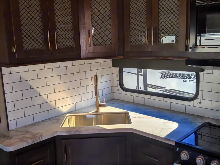 Modern RV rental kitchen: dark mesh-front cabinets, white subway tile, sink, stovetop, & microwave.. Grand Design Momentum Toy Hauler 2022