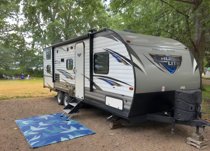 Modern RV rental travel trailer at a campsite with a blue mat. People relax under trees in the background, perfect for camping.. Forest River Cruise Lite 2019