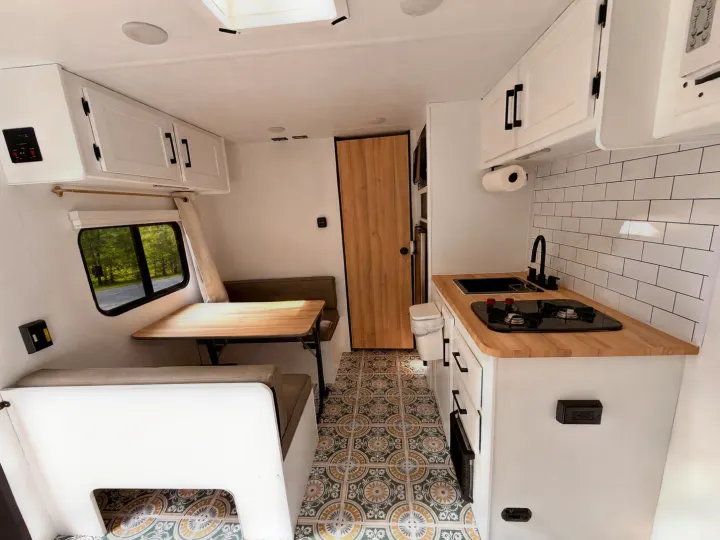 Bright RV rental interior features a kitchen with stove, sink, wood counters, a dinette, and patterned floor. Trees outside the window.. Keystone RV Coleman 2025