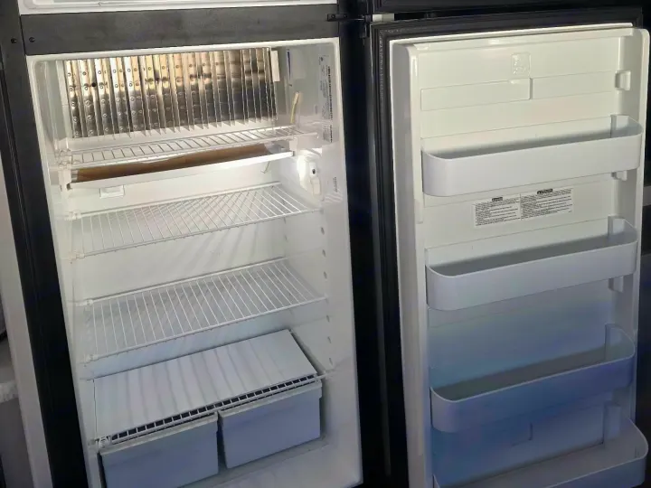 Empty RV rental refrigerator, showing the freezer with metal fins and the fresh food section with shelves and door bins, ready for use.. Keystone RV Bullet Ultra Lite 2021