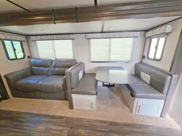 Modern RV rental interior: dark gray reclining sofa, dinette with storage, and a dining table on carpeted floor.. Keystone RV Bullet Ultra Lite 2021