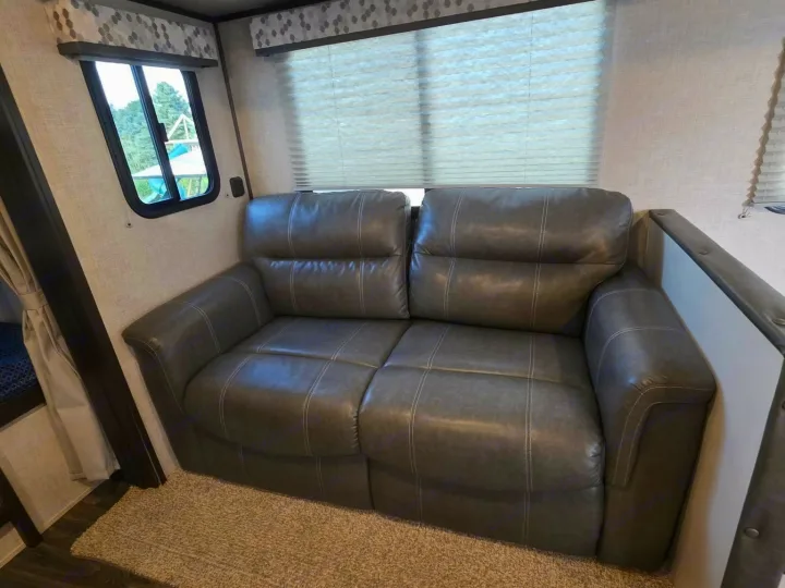 Cozy two-seater recliner sofa with white stitching in an RV rental interior, next to a window, providing comfortable seating.. Keystone RV Bullet Ultra Lite 2021