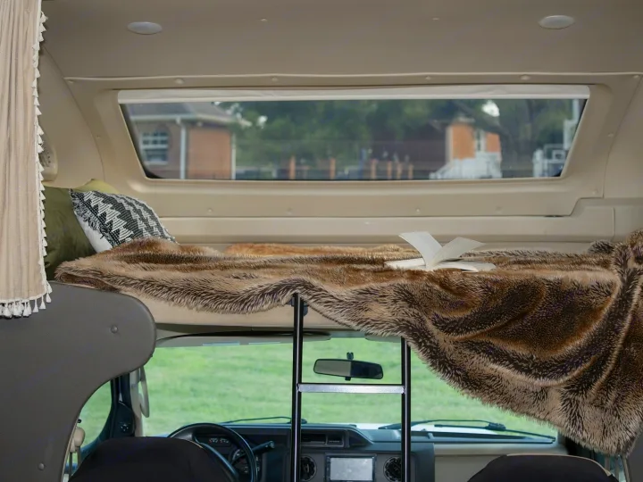 Cozy RV rental bunk bed with faux fur blanket, open book, and ladder. Through the windshield, a green landscape is visible.. Entegra Coach Odyssey 2020