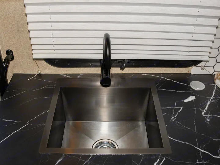 Modern RV rental kitchen with black marble-look countertop, dark metal sink, gooseneck faucet, and white pleated window blinds.. Entegra Coach Odyssey 2020