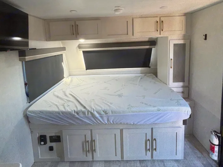 This RV rental bedroom features a comfortable bed, ample storage cabinets above and below, dark window blinds, and a TV.. No Boundaries 18 2025
