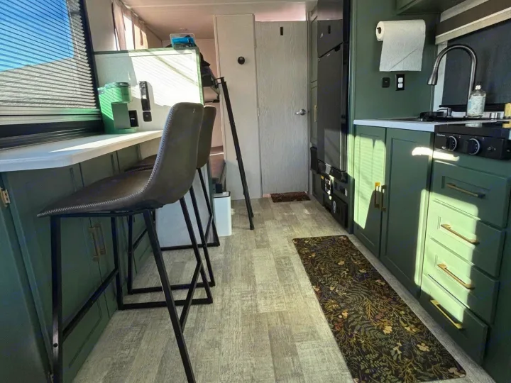 Modern RV rental kitchen featuring green cabinets, bar seating, wood-look flooring, and ample natural light through a large window.. No Boundaries 18 2025