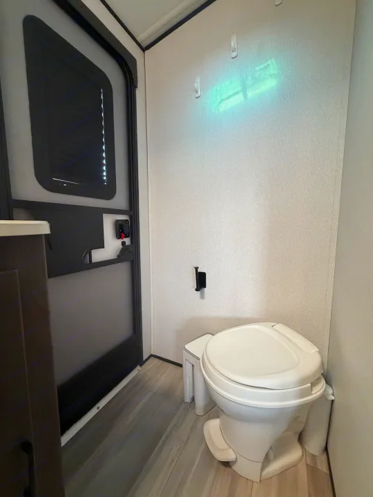 Inside a compact RV rental bathroom: white toilet, small trash bin, toilet paper holder, and door with window.. Cruiser RV MPG Ultra Lite 2021