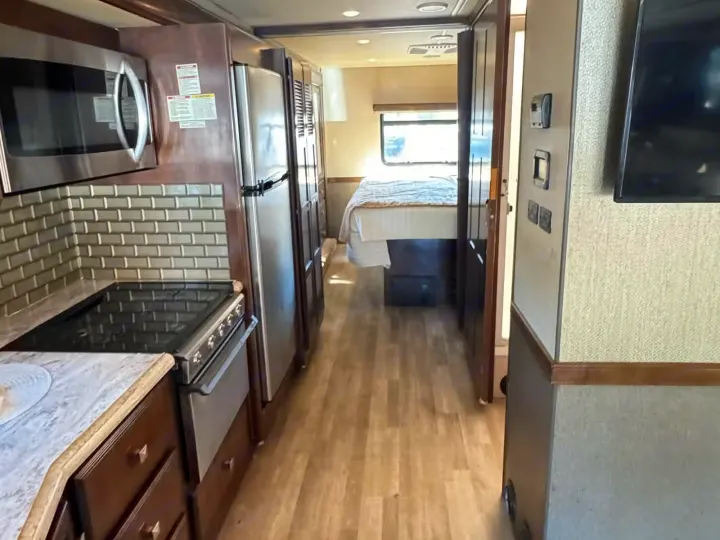 Inside this rv rental, a well-equipped kitchen with stainless steel appliances leads to a private bedroom. Light wood flooring throughout.. Holiday Rambler Admiral 2019