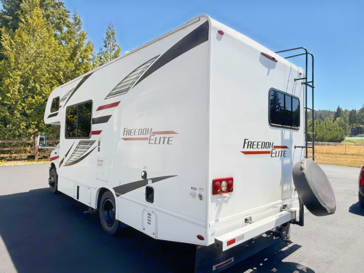 A white Freedom Elite RV rental, visible from the rear side, features black/red decals, a rear ladder, and a spare tire.. Thor Freedom Elite 2020
