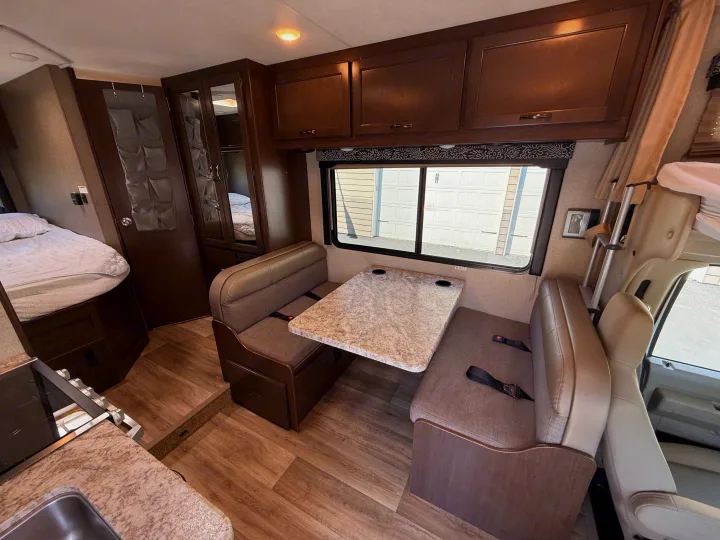 RV rental interior showing a dining area with table, kitchen, and bed. Overhead cabinets, window, and wood-look flooring.. Thor Freedom Elite 2020