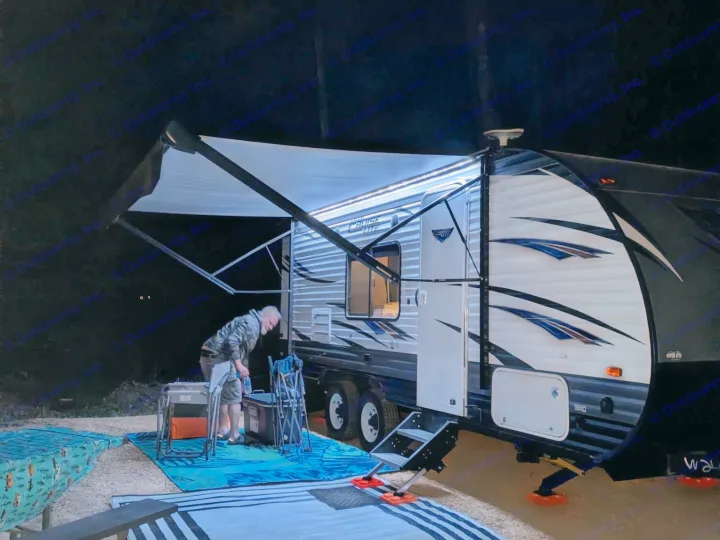 Illuminated RV rental travel trailer at night. Man bends near cooler & chairs on outdoor rugs. Awning extended.. Forest River Salem Cruise Lite 2018