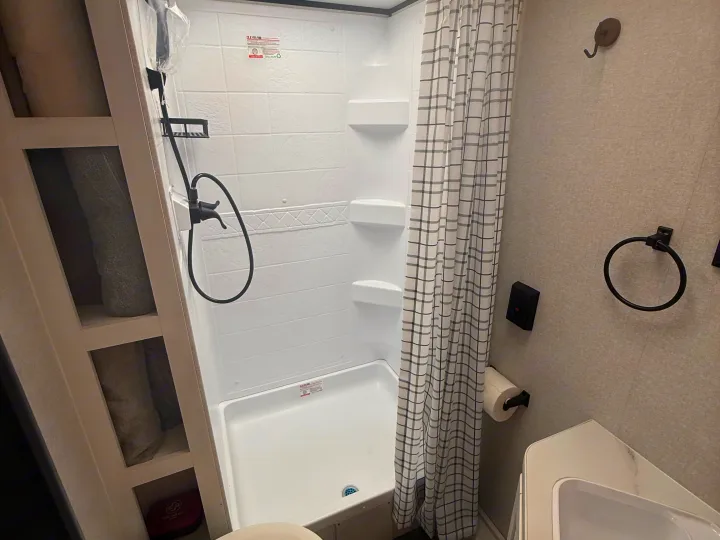 Compact RV rental bathroom features a white shower, plaid curtain, built-in shelving, toilet, and sink area, designed for travel.. Jayco Jay Flight SLX 2026