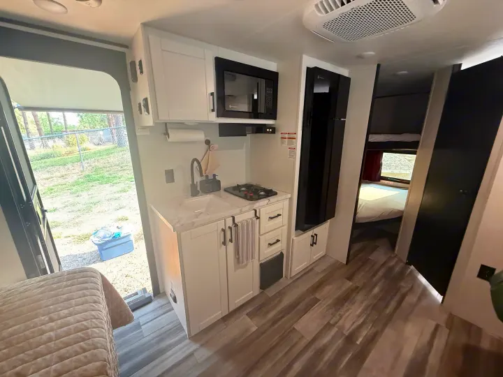Bright RV rental kitchen with white cabinets, sink, two-burner stove, microwave, fridge, & wood floor. Door opens to grass.. Jayco Jay Flight SLX 2026