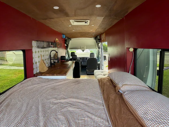 Cozy RV rental interior: a bed with brown gingham bedding, kitchen area, red walls, wooden ceiling, and front van seats.. Ford Transit 2021