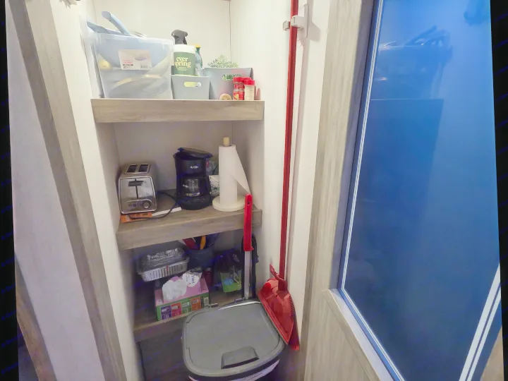 Organized RV rental interior. Shelves hold kitchen essentials like a toaster, coffee maker, and cleaning supplies. A broom and trash can are nearby.. Forest River Cherokee Alpha Wolf 2022