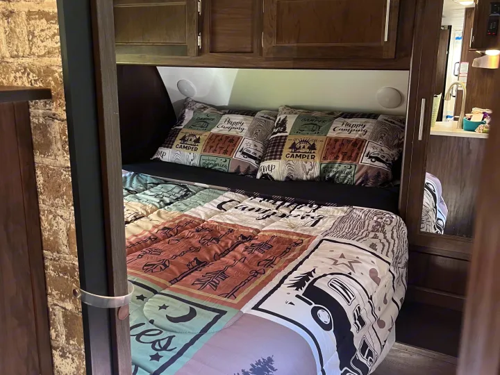 Cozy RV rental sleeping area: bed with "Happy Camping" pillows and a camping-themed comforter. Wooden cabinets and kitchen reflection.. Forest River Cherokee Grey Wolf 2018