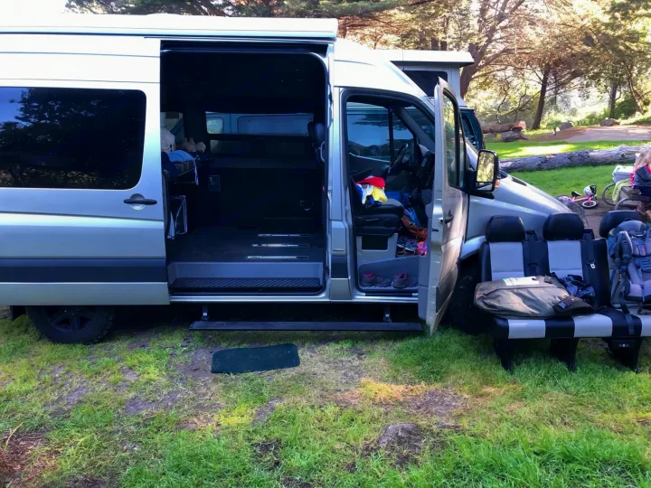 Adventure-ready RV rental. Silver camper van with open doors, showing interior bed, gear, and detached seats.. Mercedes-Benz Sprinter 2013