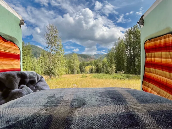 Peaceful RV rental: A cozy bed offers stunning views of mountains, forests, and a sunlit field on a cloudy day.. Ford Transit 350 High Roof EXT 2020