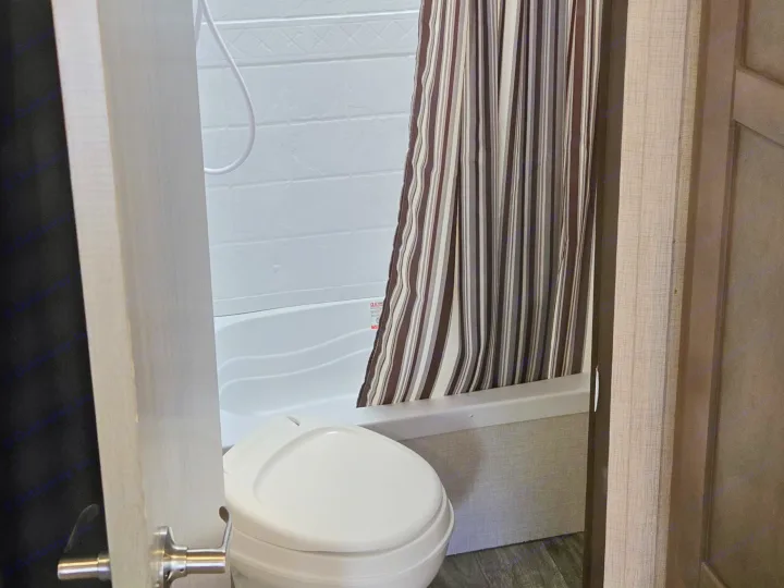This RV rental's compact bathroom features a white toilet, shower/tub with striped curtain, and wood-look flooring.. Crossroads RV Sunset Trail Super Lite 2022