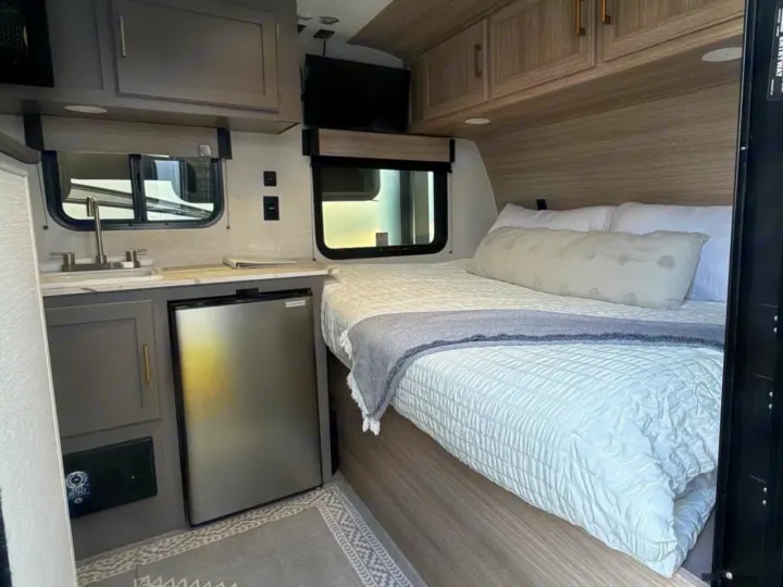 This RV rental features a compact kitchen with a sink, mini-fridge, and a cozy bed area with overhead cabinets.. KZ Sportsmen Classic 130BH 2026