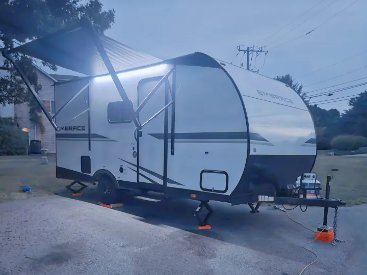 This RV rental, a sleek travel trailer with bright awning lights, is set up for a cozy evening, stabilized on leveling blocks.. Cruiser RV Embrace 2025