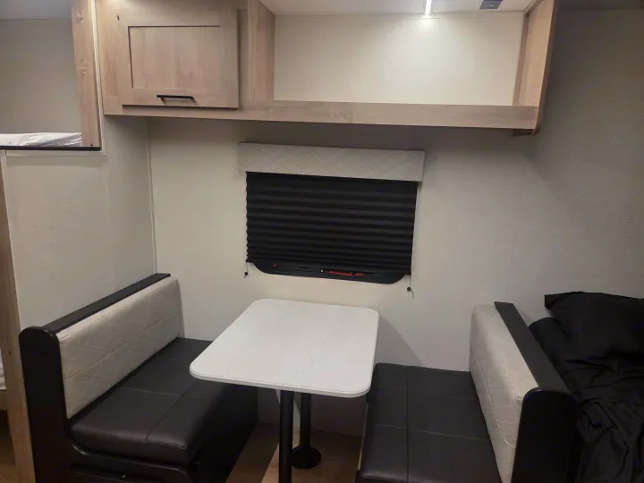 Interior of an RV rental showcasing a cozy dinette with dark benches, a white table, overhead cabinets, and a window blind.. Cruiser RV Embrace 2025