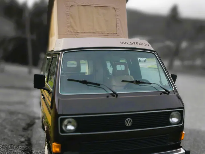 A brown Volkswagen Westfalia pop-top camper van with its roof extended, perfect for an RV rental, is parked outdoors.. Volkswagen Westfalia 1985